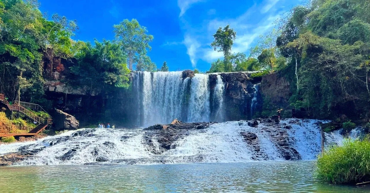 Bousra Waterfall in Mondulkiri, Cambodia: water falling over dark rock into a pool, framed by forest and blue sky.